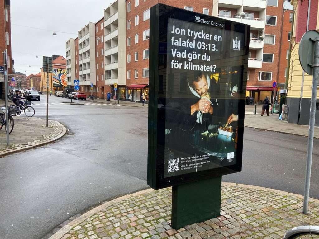 Bild från ett bostadsområde där en storskärm visar en bild på en person som äter falafel. Texten lyder "Jon trycker en falafel 03:13. Vad gör du för klimatet?"