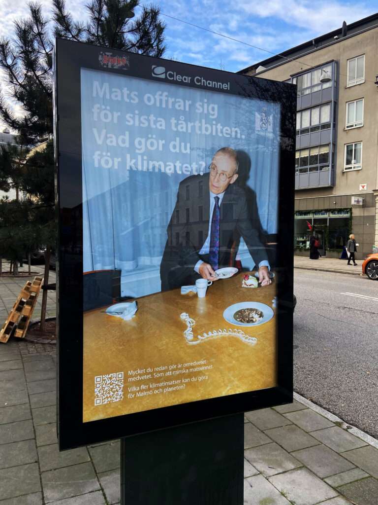 En bild på ett stortryck i ett bostadsområde. På trycket tar en man en tårtbit. Bildtexten lyder "Mats offrar sig för sista tårtbiten. Vad gör du för klimatet?"